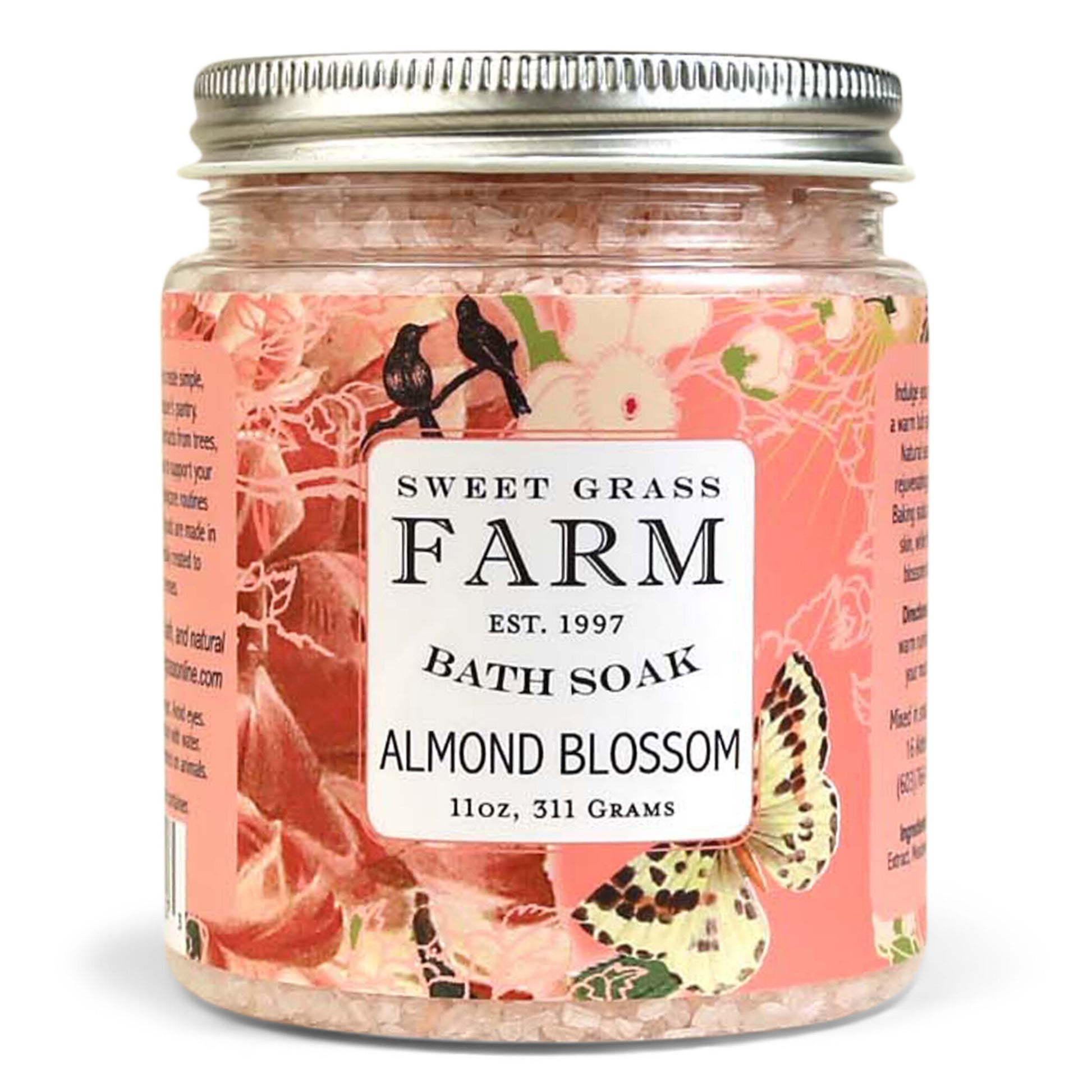 A jar of Sweet Grass Farm bath soak with a label that reads 'BATH SOAK ALMOND BLOSSOM 11oz, 311 Grams'. The jar is filled with pink bath salts.