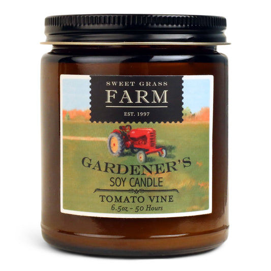 A jar candle with a label that includes a red tractor and text, indicating the brand 'SWEET GRASS FARM' and the product name 'GARDENER'S SOY CANDLE TOMATO VINE'. The jar is transparent and is made of glass.